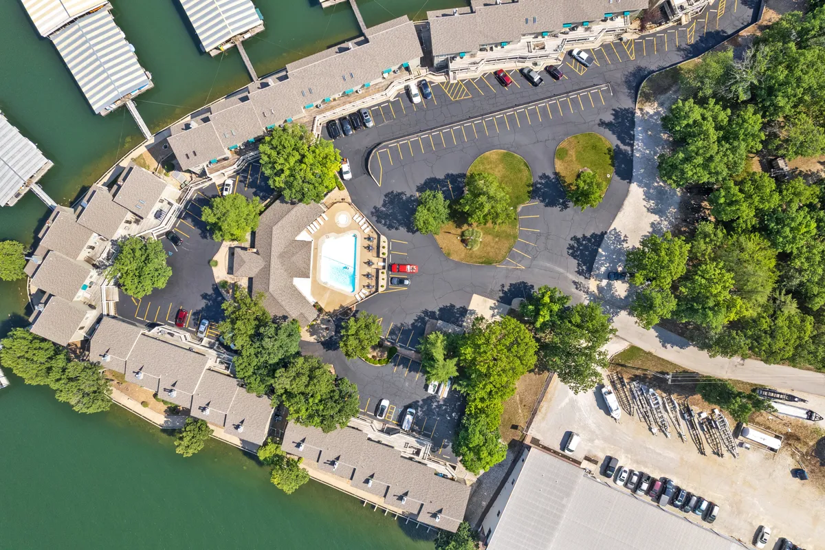 A scenic view encompassing a swimming pool, dock area, parking lot, and surrounding trees. The lake is visible in the background, enhancing the sense of lakeside tranquility. The communal spaces suggest opportunities for swimming, boating, and water activities, as well as sunset views.