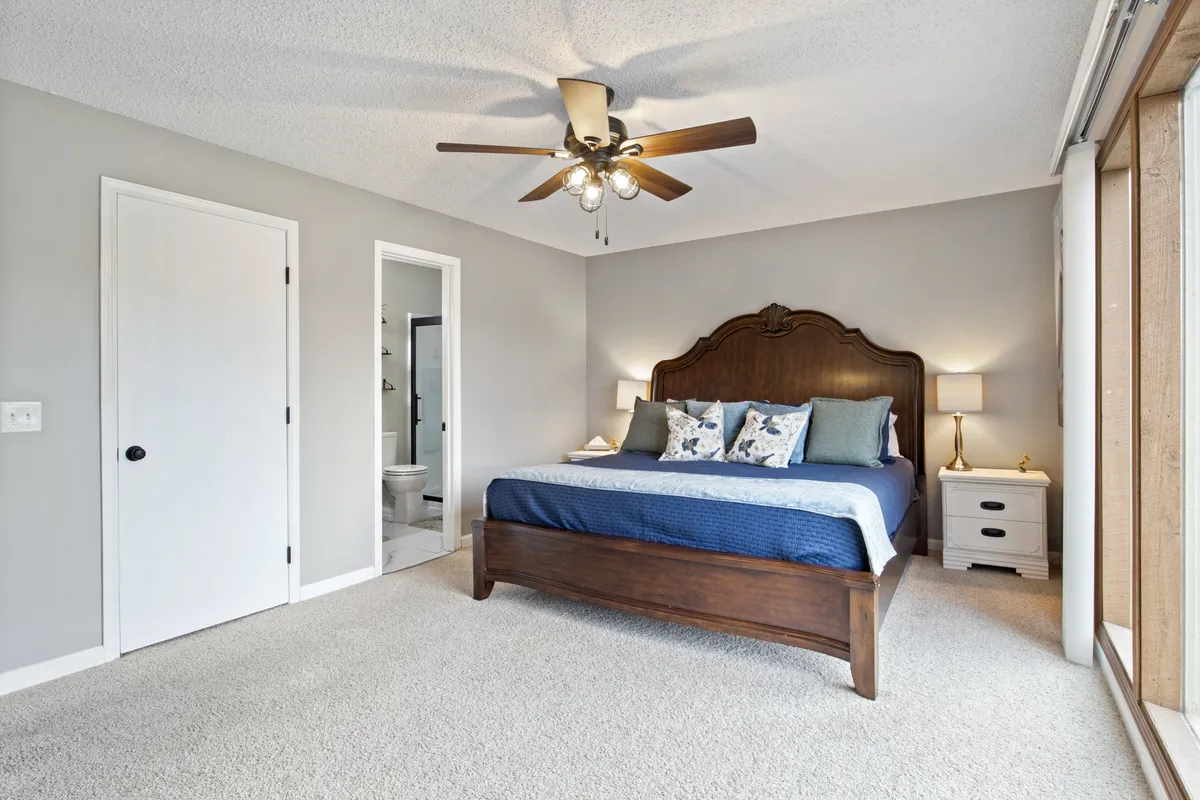 A serene bedroom with a well-made bed accented by decorative pillows, a nightstand with a warm lamp, and a ceiling fan overhead. The space exudes comfort and an inviting atmosphere for relaxation or reading.
