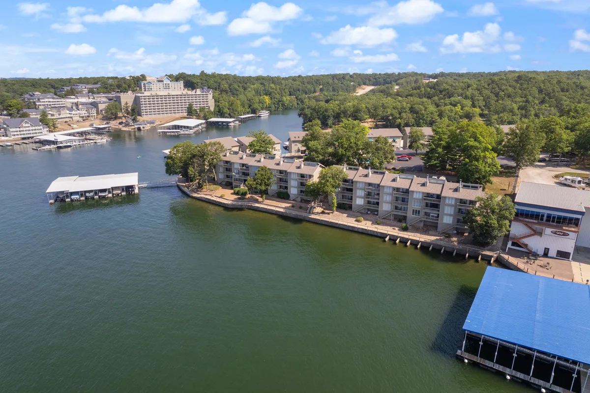 A vibrant exterior scene featuring waterfront buildings, a community dock, lush greenery, and several boats moored along the dock. The image conveys the essence of lakeside living, with easy access to boating and a shared community atmosphere.