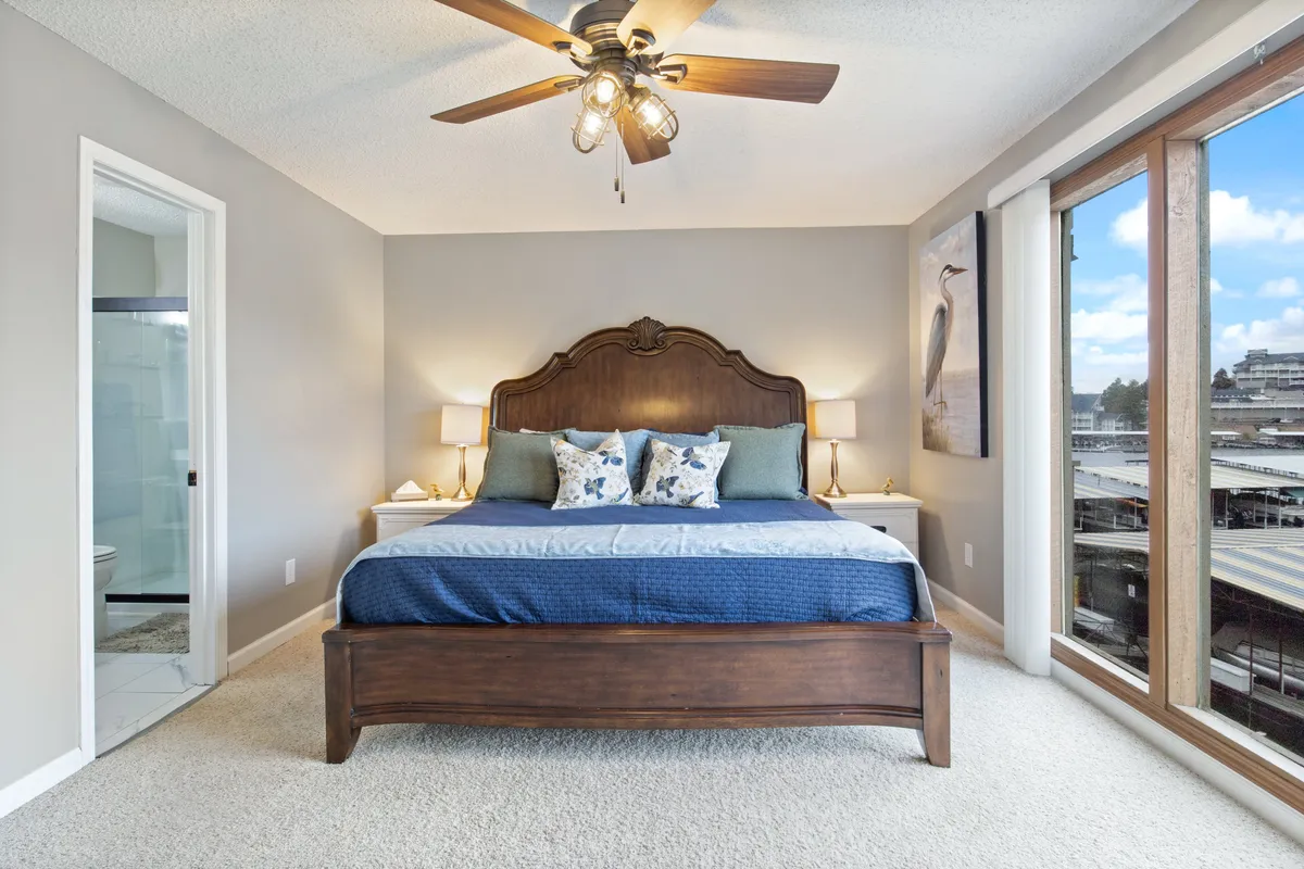 This image showcases a king-sized bed adorned with decorative pillows, flanked by wooden bedside tables, and bathed in natural light streaming through large windows. The ambiance is serene, emphasizing restful comfort and a sophisticated, uncluttered aesthetic.