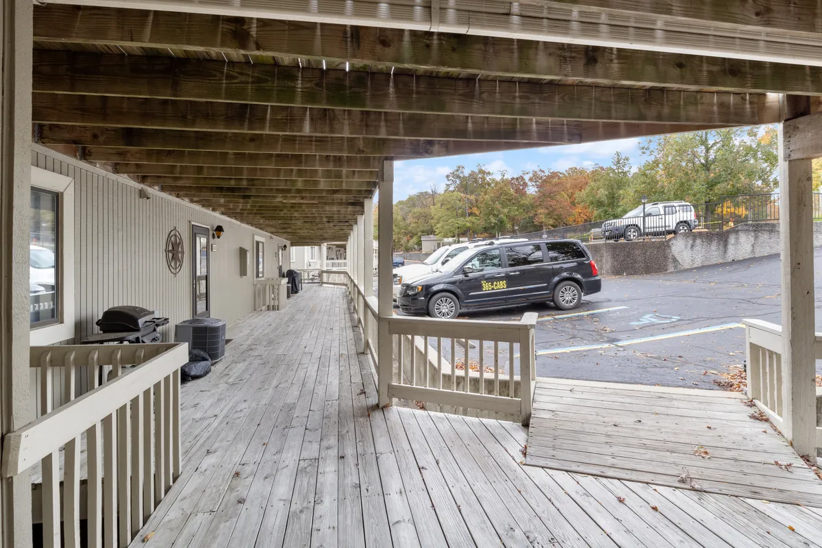 A wooden walkway leads past the parking area with visible vehicles, inviting guests to stroll toward their home base. The relaxed vibe, with socializing on the porch and the proximity to vehicles, reinforces convenience for launching day trips to local attractions or the lake.