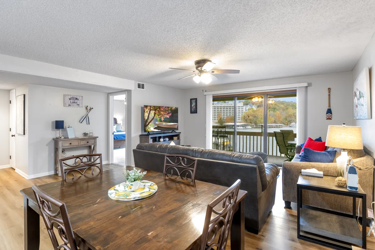 A comfortable dining table and living room seating area are arranged near a large window, offering ample natural light and scenic lake views. The space is ideal for family dinners, relaxing with friends, and enjoying the outside scenery from within. The arrangement is inviting and emphasizes both comfort and sociability.