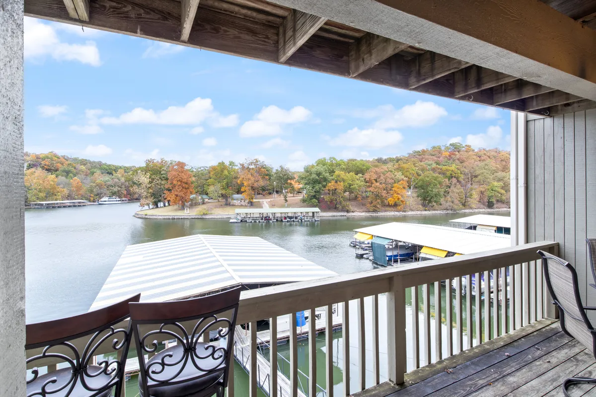 The image showcases a peaceful lake view framed by vibrant autumn foliage, with charming boathouses nestled along the shore. A sturdy deck railing suggests a spacious outdoor area perfect for relaxing, while the calm water reflects the rich colors of fall. The scene evokes possibilities for boating, fishing, and tranquil moments spent outdoors.